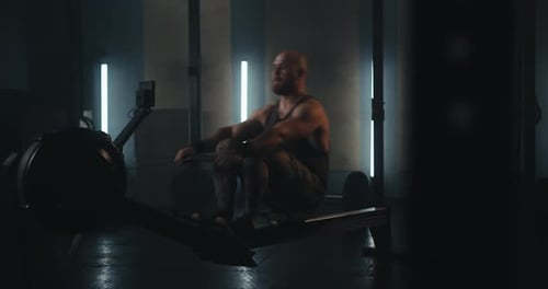 Muscular Bearded Man in a Tank Top Working Out on a Rowing Machine in a Modern Gym