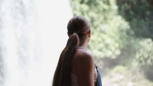 Eine junge Frau in einem Jeans-Overall bewundert den Wasserfall in einem Gebirgsfluss im Wald.