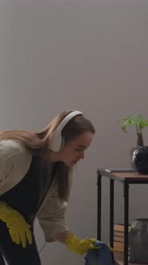 Young Woman Wearing Headphones and Rubber Gloves Happily Dusting and Wiping Living Room Surfaces