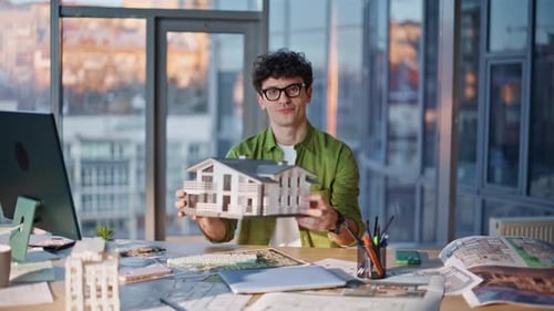 Smiling architect presents a house model at desk