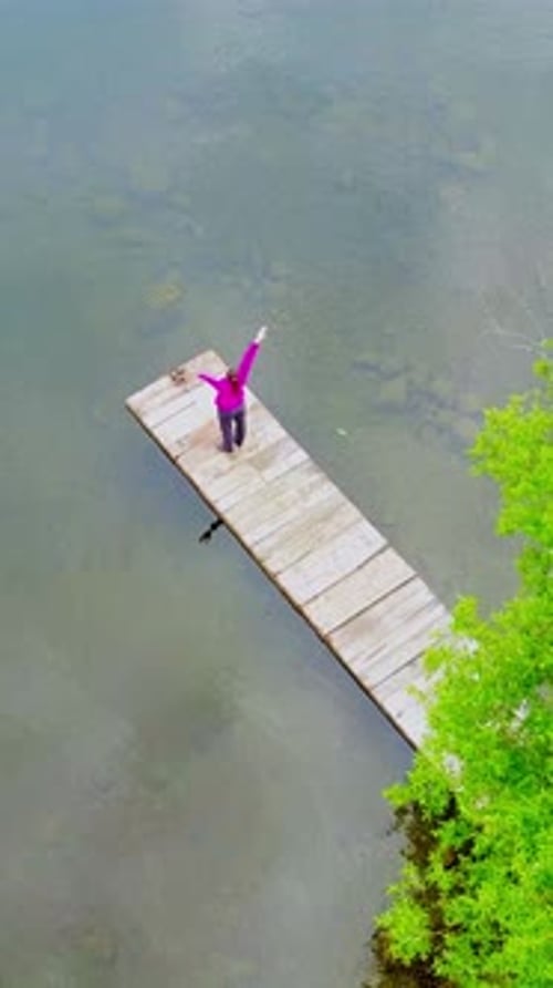 Woman Standing on Wooden Dock By Clear Lake Surrounded By Lush Greenery in Serene Nature Setting