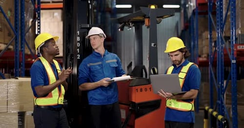 Multiracial male workers wearing safety vests discussing job tasks in warehouse as uses hand gesture