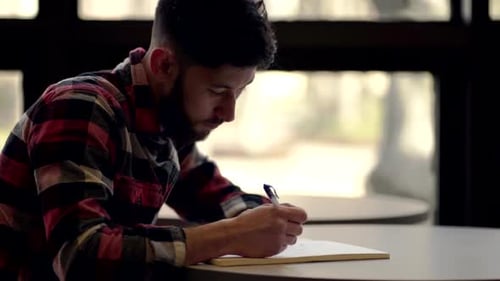 A Young Man Wearing Red Long Sleeve Writing On His Notes in the Table - Close-Up Shot