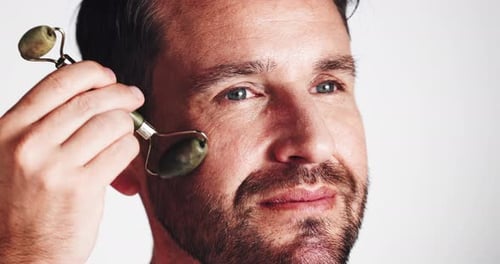 Man, skincare and jade roller in studio with cosmetics, routine and process for anti aging by white