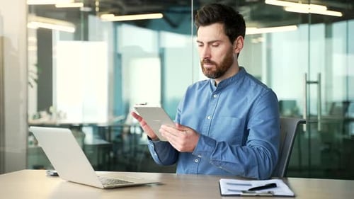 Handsome bearded businessman is using digital tablet sitting at workplace in business office.