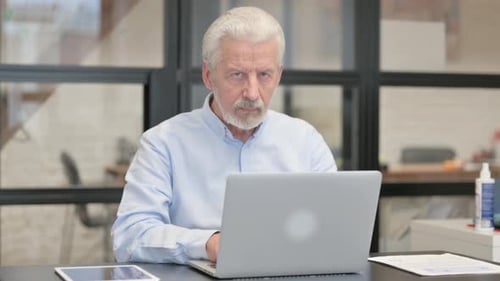 Old Businessman Looking at Camera while Working on Laptop in Office