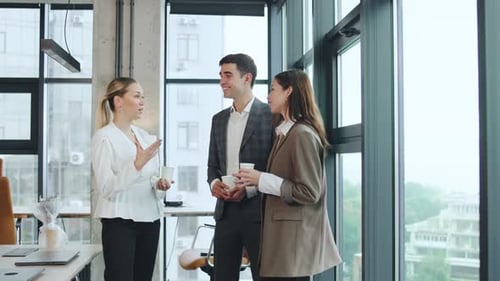 Business Meeting Takes Place Indoors with Three People Talking and Holding Drinks in a Modern Office