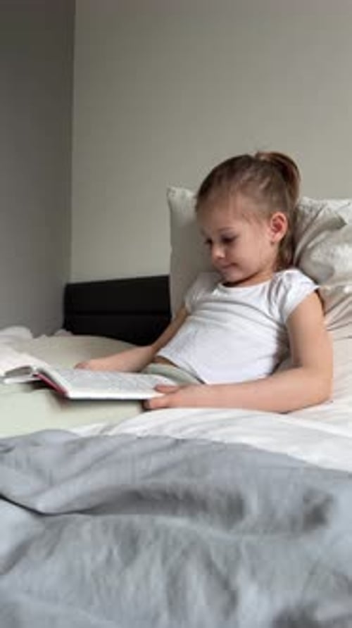 Little Girl Sitting on Her Parents Bed and Reading a Book in the Bedroom Concept of Quiet Time