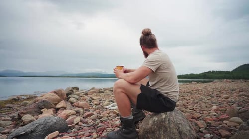 Bearded Man Hiker Sitting By A Rocky Shore Having A Cup Of Warm Drink In Anderdalen National Park, S