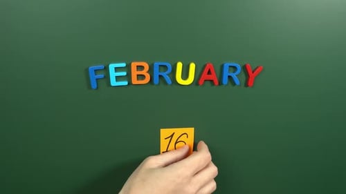 Hand sticking a sticker 16 February calendar day on school board. 16 date of February. Sixteenth