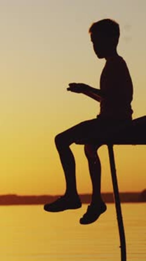 Silhouette eines Jungen, der auf einer alten Brücke am Fluss spielt. Wunderschöner Sommersonnenuntergang Vertikales Video