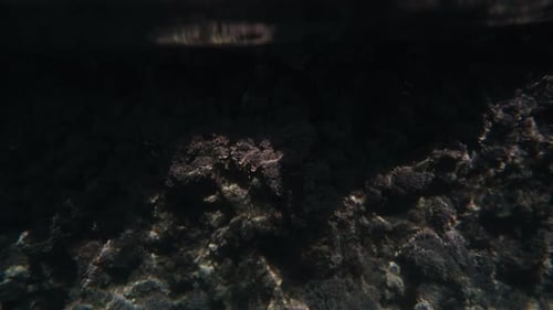 The camera at the water's edge captures the transparent sea, with rocks both on the shore.
