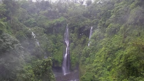 Aerial flies through jungle valley mist toward multi cascade waterfall