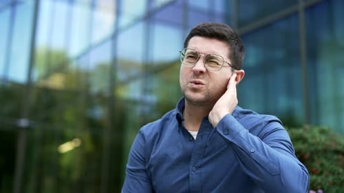 Sick businessman suffers from ear pain sitting on a bench on the street. Upset male touches his ear