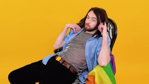 Gay Man Dressed in Mesh Tshirt Sits in a Chair with a Multicolored Flag