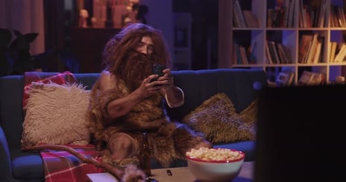 Furry Bearded Human Ancestor in Animal Fur Sitting on Couch Watching TV Using Mobile Phone
