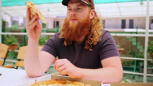 Man squinting with pleasure bites off a piece of juicy cheese pizza sitting in a street cafe