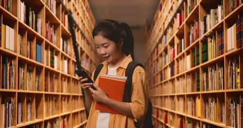 Asian Woman Student With A Backpack Smiling And Using A Smartphone While Standing In The Library