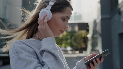 Woman Adjusts Headphones While Using Smartphone Outdoors