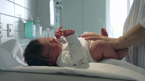 Newborn baby being dressed in a white outfit by a parent, lying on a hospital changing table,