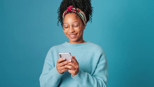 Young smiling woman having fun with mobile phone on blue background in studio