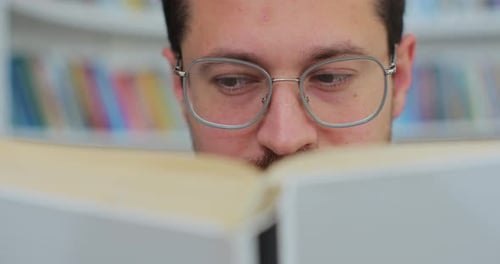 Nahaufnahme: Ein Student mit Brille verbringt seine Zeit in der Bibliothek, untersucht ein Buch und liest