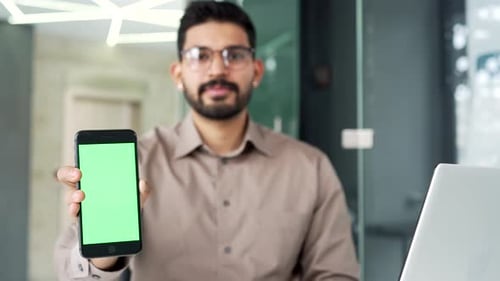 Smartphone with green screen showing happy businessman sitting at workplace in business office.
