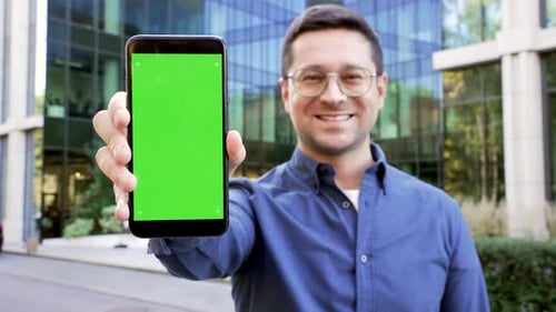 Happy handsome businessman showing mobile phone with green screen standing on street near a business