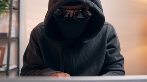A hacker in a dark hoodie uses a laptop.