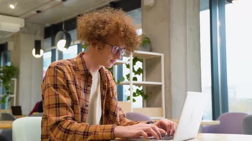 Tired Disappointed Programmer Sitting at a Table in a Cafe and Working on a Laptop Wears Yellow