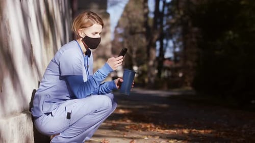 Woman in scrubs sitting using phone with coffee
