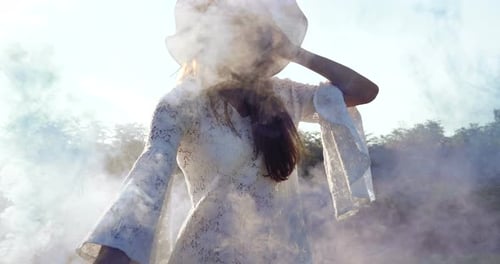On a sunny day, a woman walks across the field with a colored smoke flare in a white dress against