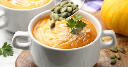 Adding seeds into tasty pumpkin cream soup in bowl on table, closeup