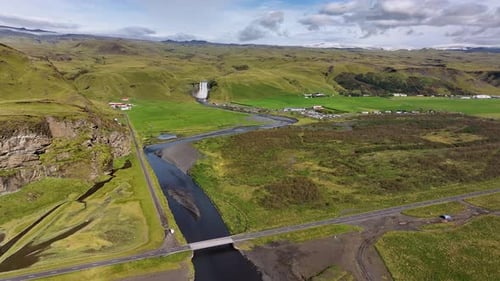Drone shot approaching Skógafoss waterfall in Iceland over green landscape, with river, road, and