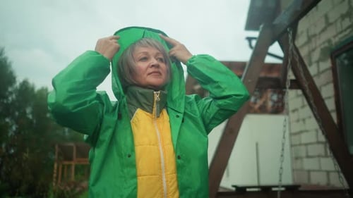 Middleaged Woman Adjusts Green Raincoat Hood Near Swing Bench in Outdoor Setting