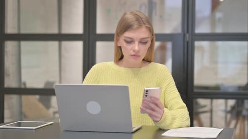 Young Woman Using Phone and Laptop at Desk