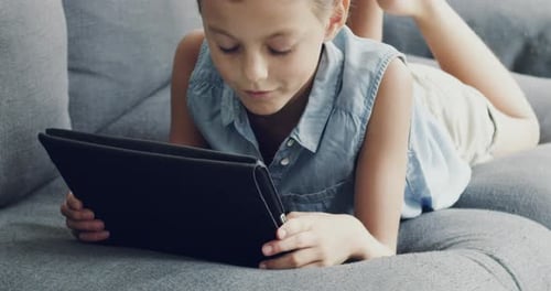 4k video footage of a little girl lying on a sofa at home using a digital tablet