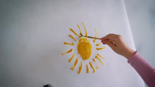Close-up of a child's hand with a brush painting a yellow sun on a sheet of paper.
