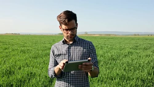 Porträt eines Landwirts, der auf einem jungen Weizenfeld steht, eine Tablette in der Hand hält und die Ernte untersucht.