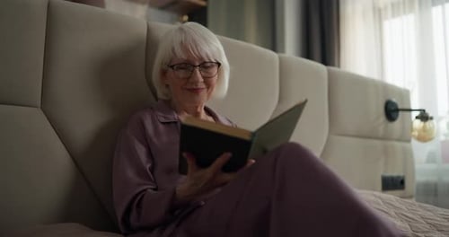 Senior Woman in Pajamas with Glasses Reading Her Favorite Book and Smiling While Relaxing on Bed in