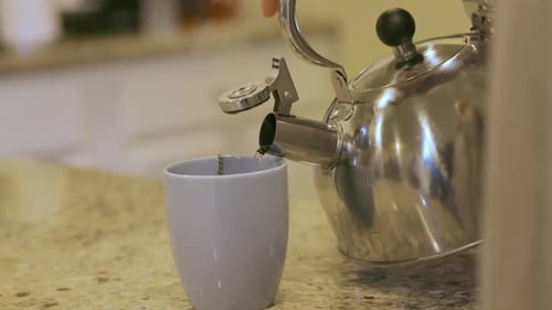 Metal teapot pouring green tea into cup. White mug and stainless steel teapot or tea kettle.