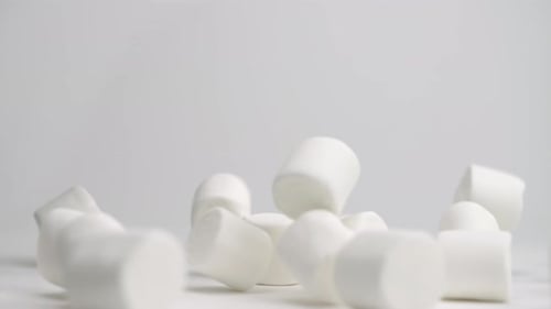 White squishy jumbo marshmallows falling onto white table top and bouncing around into a pile in slo