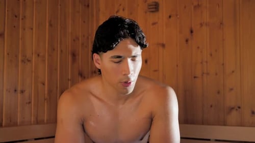 Homme asiatique assis sur un banc avec une serviette dans le sauna, voyant la fenêtre, se retournant, s'essuyant le visage, respirant lentement