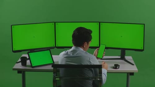 Asian Man Using Mobile Phone And Multiple Computer Monitor With Mock Up Green Screen In Green Screen