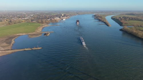 Aerial view of two large cargo ships navigating a wide river, with grassy banks, small peninsulas