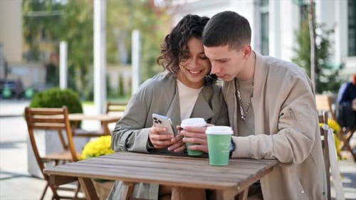 A happy couple outdoors near a cafe. Looking in the phone, smiling, talking, coffee. Autumn atmosphe