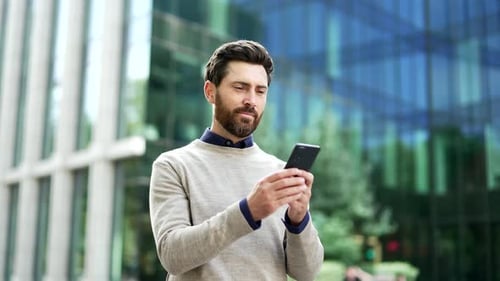 Happy businessman is using a phone standing on the street near a business office building.