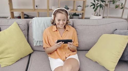 Top view, Asian woman on sofa and headphones with cellphone, streaming music and funny videos