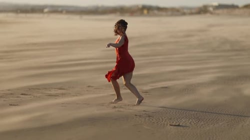 Graceful Young Woman in Red Dress Dancing on Sandy Beach in the Wind