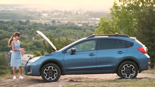 Puzzled Young Female Driver Standing Near a Broken Car with Open Up Hood Inspecting Her Vehicle